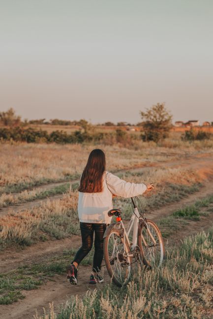 Back view of faceless lady with dark hair rolling bicycle on grassy road in countryside in summer evening in nature