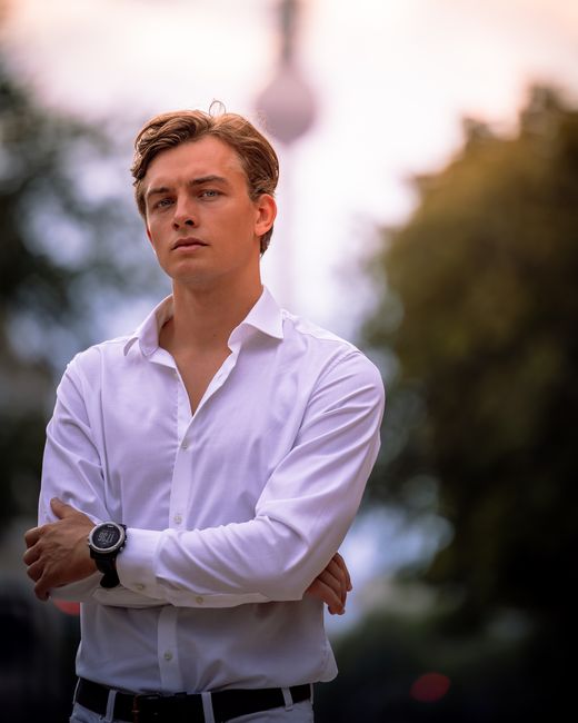 Portrait of a young man in a white shirt standing confidently in an outdoor park setting.