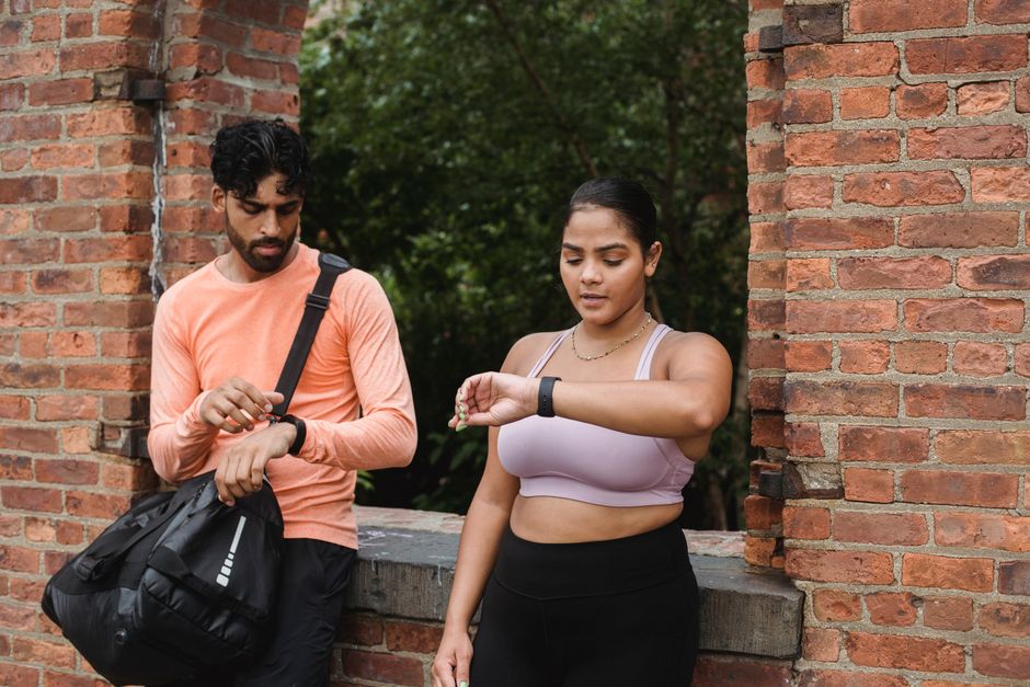 Two adults in sportswear checking their smartwatches by a brick wall outdoors.