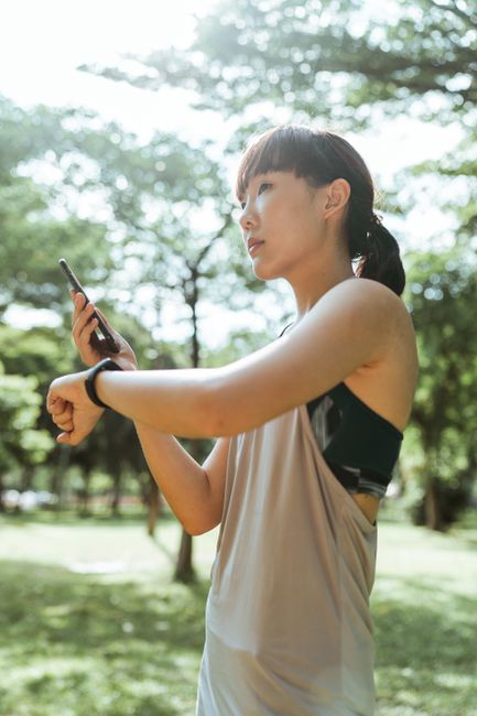 From below of Asian female wearing sportswear standing in park and using smart watch with mobile phone