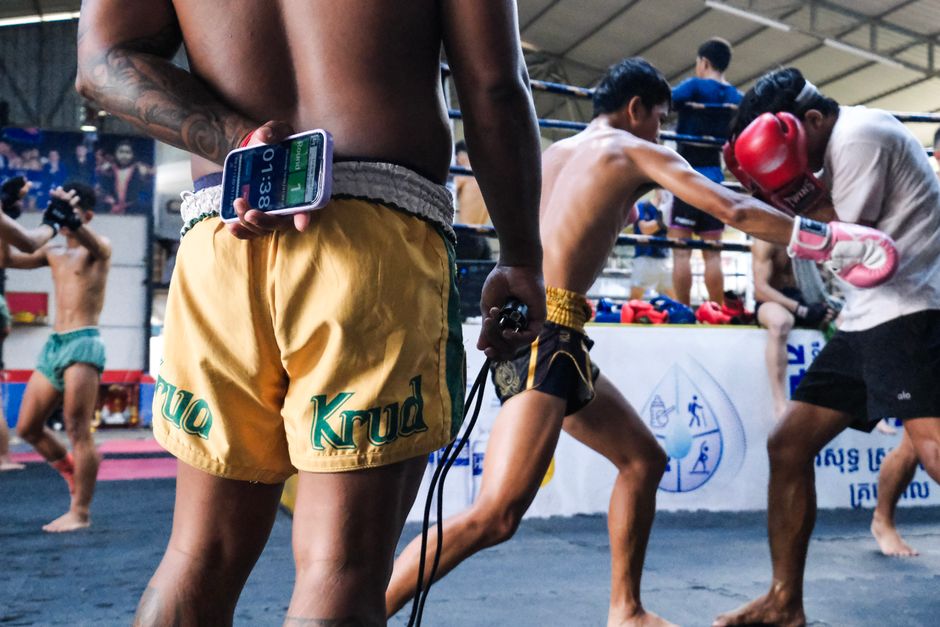 Dynamic scene of Muay Thai fighters training in a gym with a coach timing the session.
