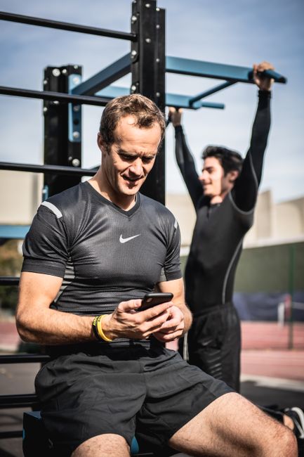 Two men working out on outdoor fitness equipment, one using smartphone.
