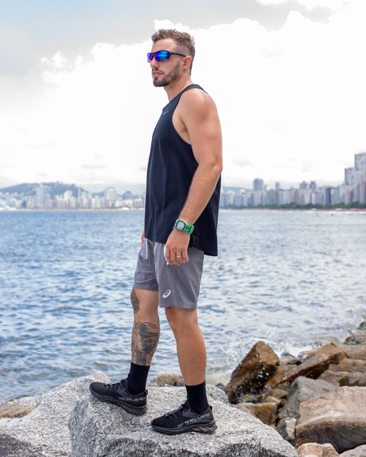 Athletic man in casual sportswear standing on rocks by the sea, with a city skyline in the background.