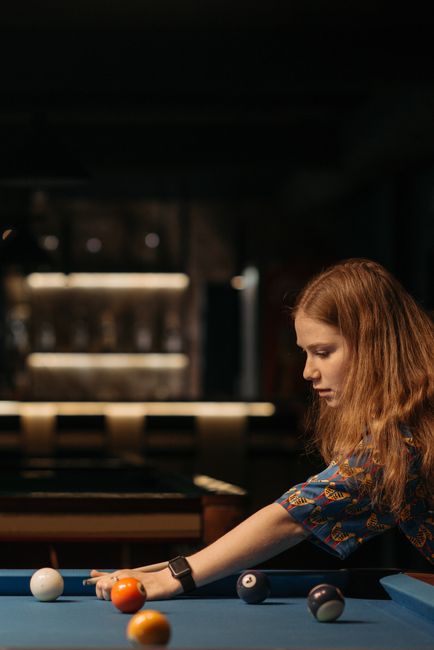 Focused woman playing billiards, aiming at cue ball on a blue table in a dimly lit indoor setting.