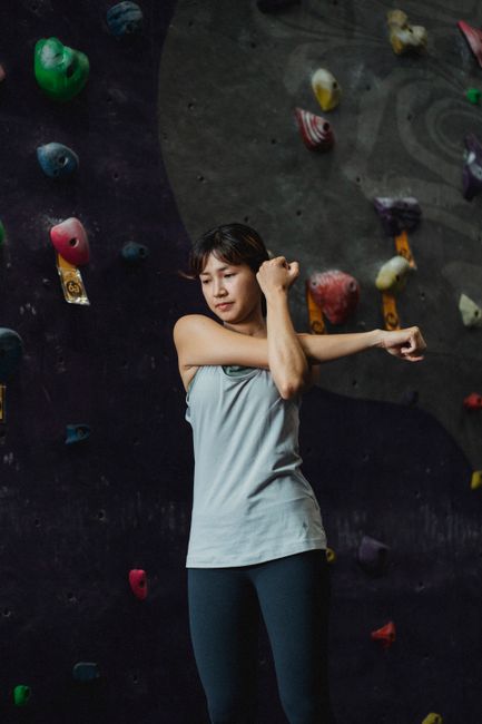Young fit ethnic female mountaineer in sportswear stretching arm and looking down during workout near artificial rock