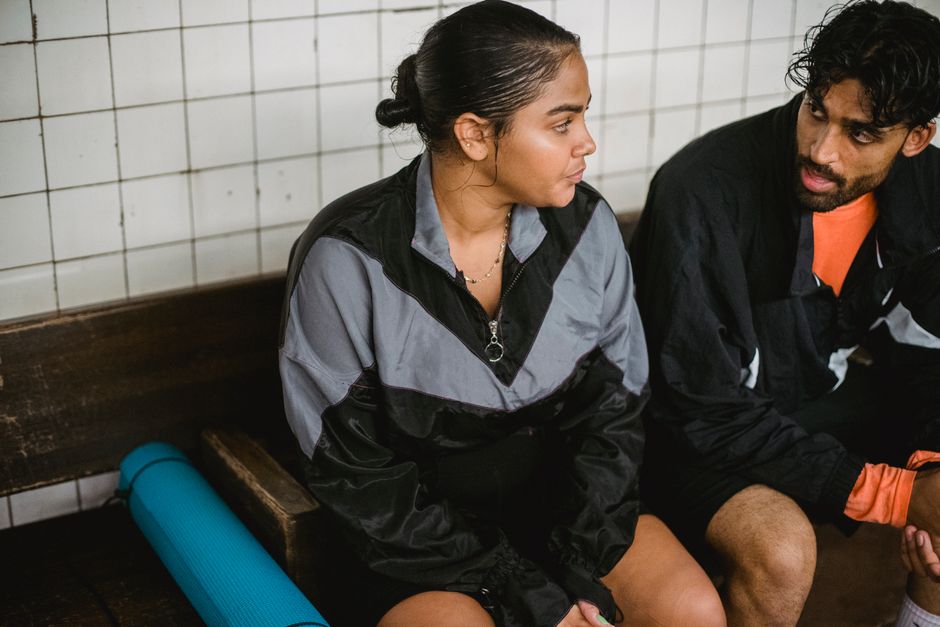 A man and woman in sportswear relax on a bench indoors post-workout, enjoying some downtime.