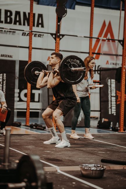 Athlete lifting barbell in indoor gym competition setting with coach nearby.