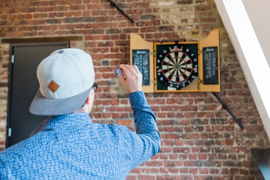 An adult man in a blue shirt playing darts indoors, aiming at a brick wall-mounted dartboard.