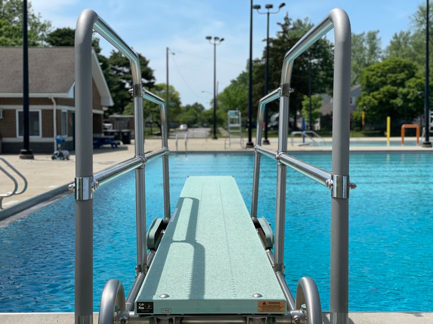 View of an empty diving board leading to a clear blue swimming pool outdoors.