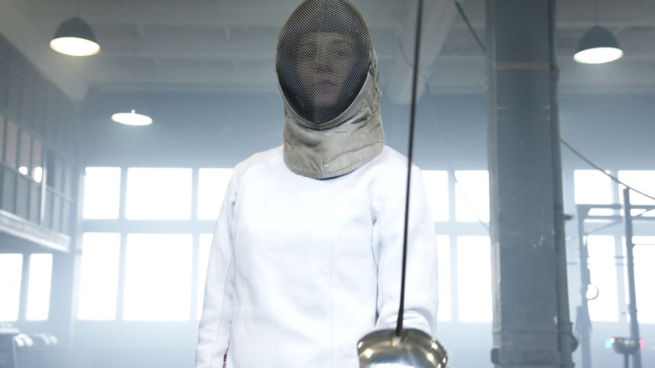 Female fencer wearing mask and gear, holding a sword in an indoor setting.