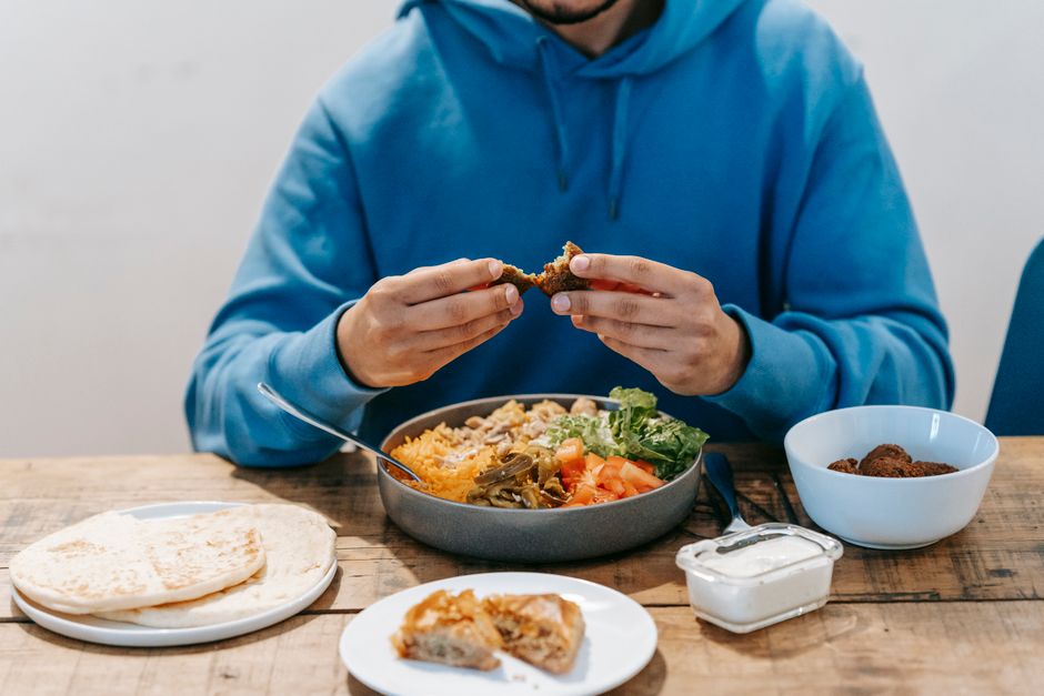 Crop unrecognizable male tasting fried meat from dish with fresh vegetables at wooden table with plates filled with food