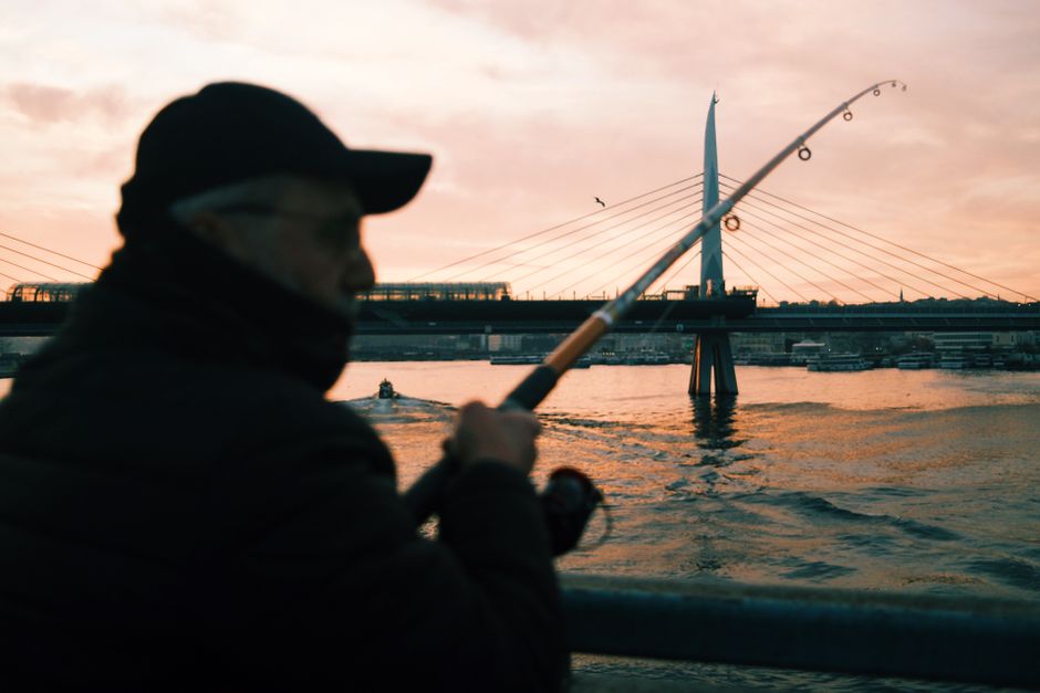Silhouette of a fisherman casting near a contemporary bridge at sunset.