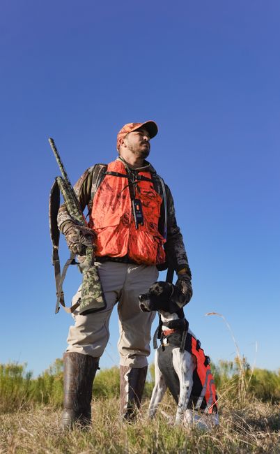 Man in hunting gear with trained pointer dog outdoors, under clear blue sky.
