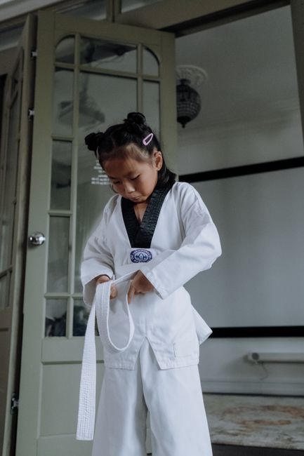 Asian girl in karategi tying her white belt indoors, focusing on her training.