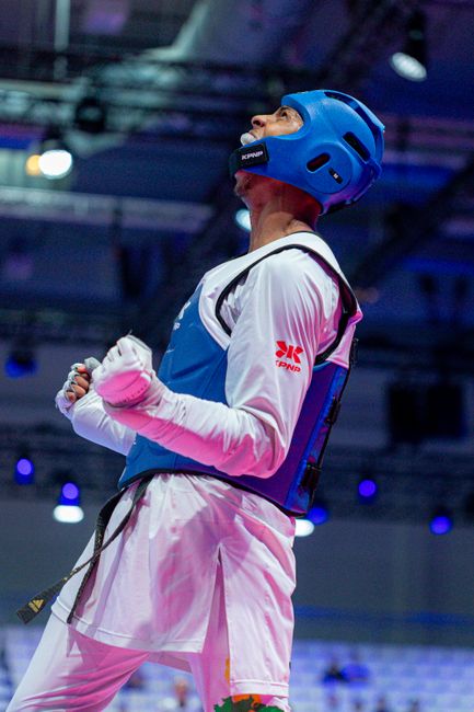 Male taekwondo athlete in blue gear celebrating victory in arena with dynamic lighting.
