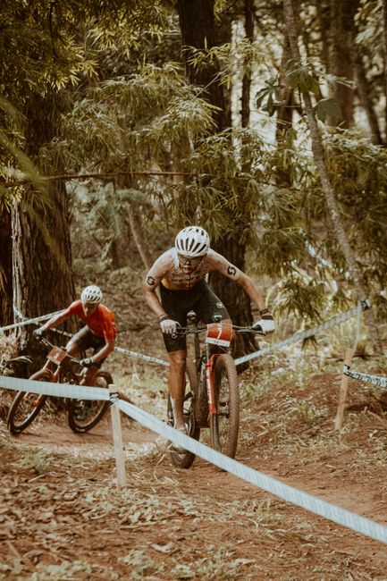 Two cyclists compete in an off-road mountain biking race through dense forest trails.