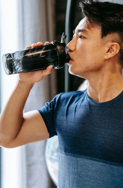 Serious ethnic man in sportswear drinking water from plastic bottle during break in fitness workout in gym
