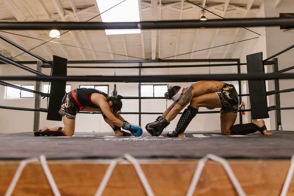Two martial artists, one male, one female, stretch inside an indoor training ring.