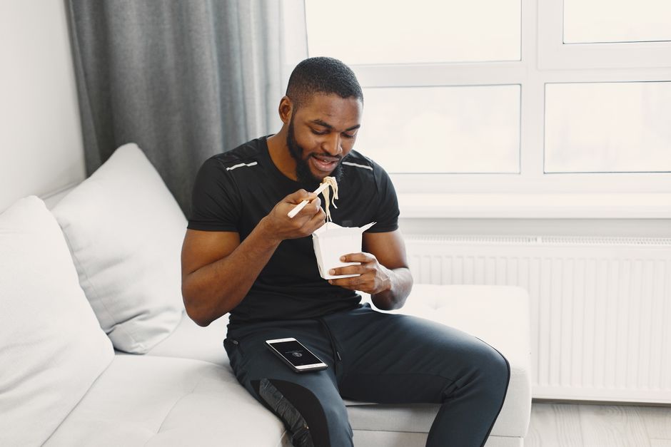 Casual man enjoying takeout with chopsticks while relaxing on a couch indoors.