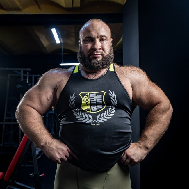 A strong bodybuilder poses confidently in a gym, showcasing muscular physique in a tank top.