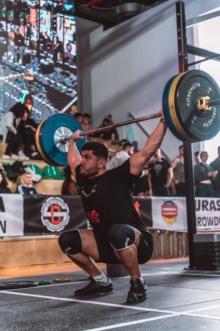 A focused athlete lifts weights at a competitive indoor event with a crowd watching.