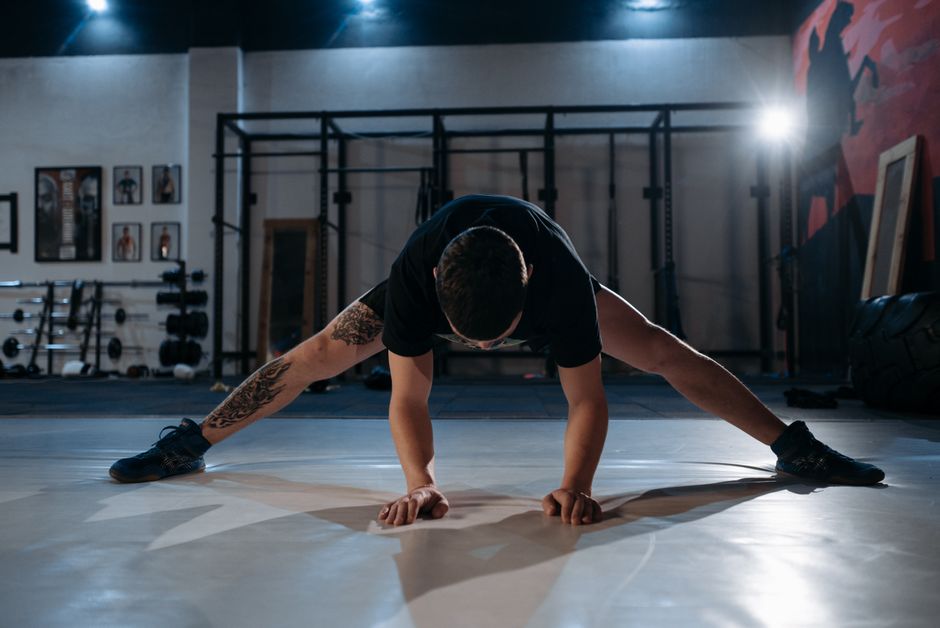 A man with a tattoo stretches on a gym floor under dramatic lighting.