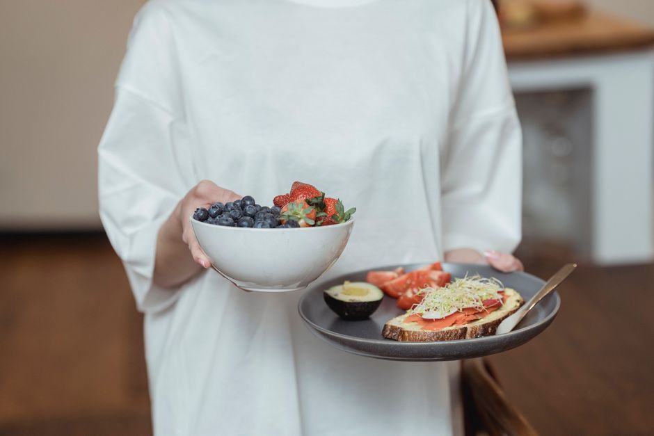 Person holding a bowl of fresh berries and a plate with avocado toast, displaying a healthy breakfast spread.