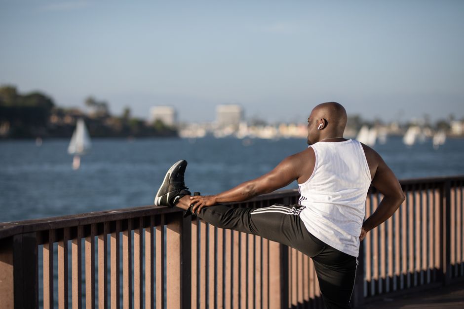 Bald man in activewear stretching against railing near water under clear sky.