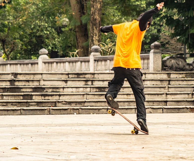 A skateboarder in an orange shirt performs tricks in a lively urban park setting.