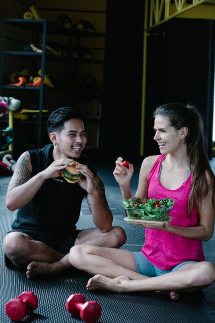 A couple laughs while sharing a salad and burger sitting on a gym floor.