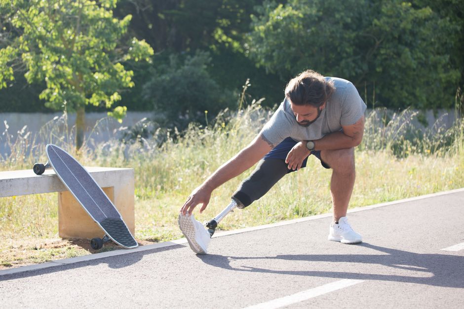Active man with prosthetic leg stretching on a pathway next to a longboard.