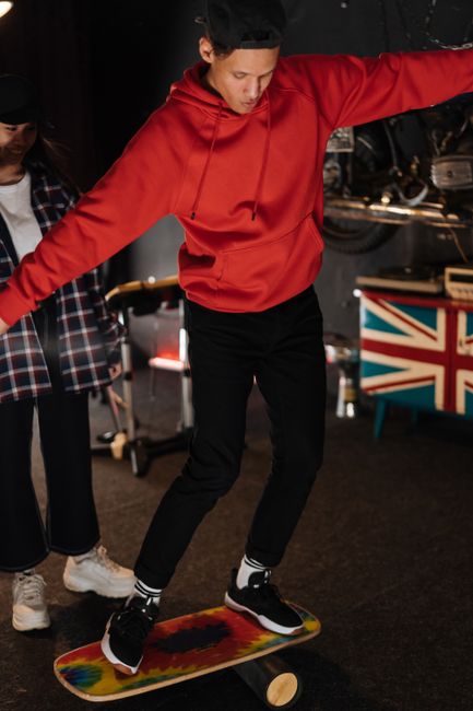 Young skater in red hoodie balancing on a skateboard indoors with a friend nearby.