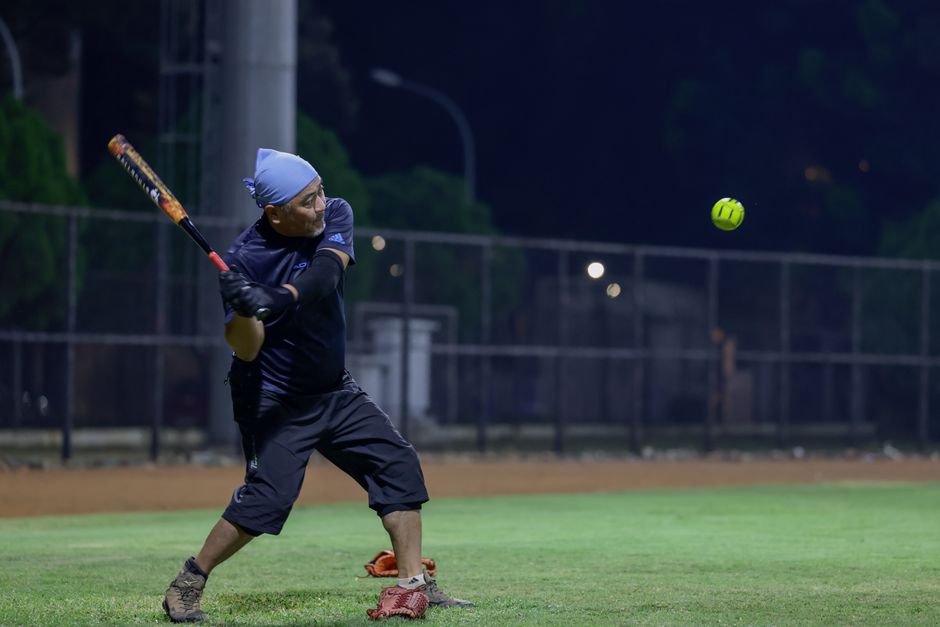Male player batting during a nighttime softball game under stadium lights.