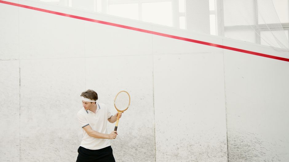Active squash player in white sportswear preparing for a serve on indoor court.