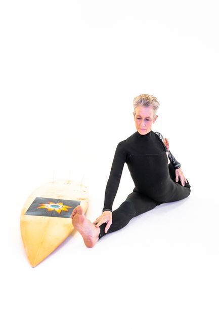 Senior woman in wetsuit stretching indoors next to a surfboard.