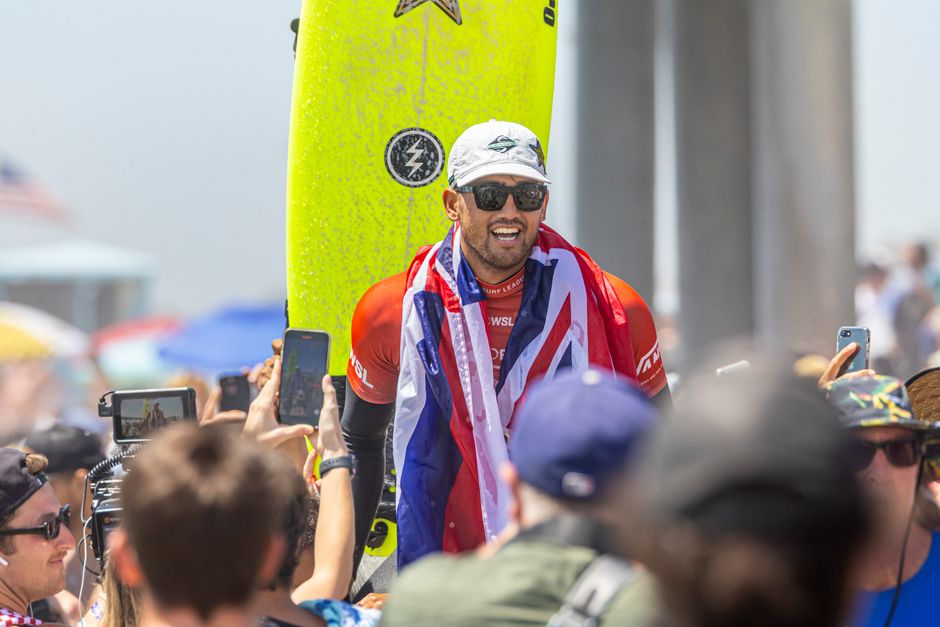 Surfing champion celebrates victory wrapped in flag, surrounded by fans at sunny beach event.