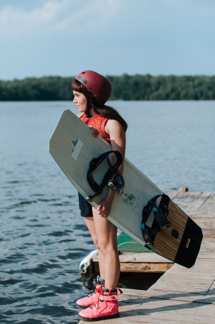 A woman in a helmet stands on a pier, holding a wakeboard, ready for an adventure on the lake.