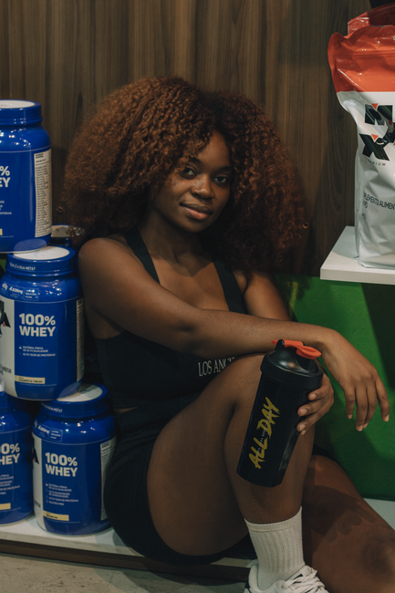 Young woman with curly hair, seated with protein containers, embodying fitness lifestyle.