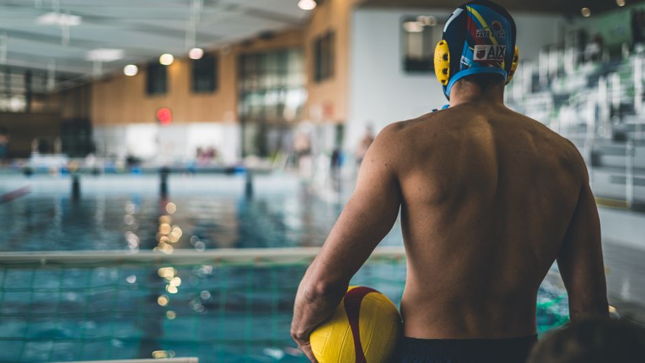 Male water polo player holding a ball, preparing for a match at an indoor pool.