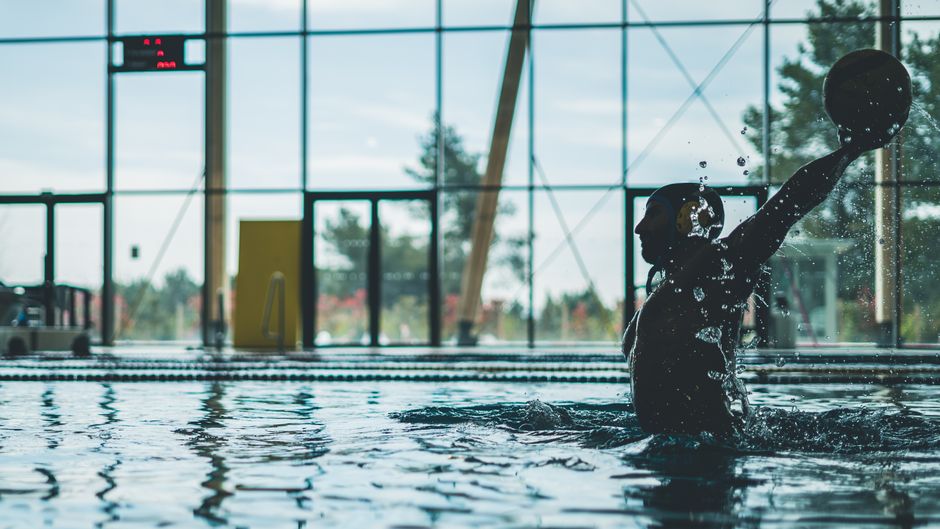 Silhouette of a water polo player mid-action with water splashes inside an indoor pool.