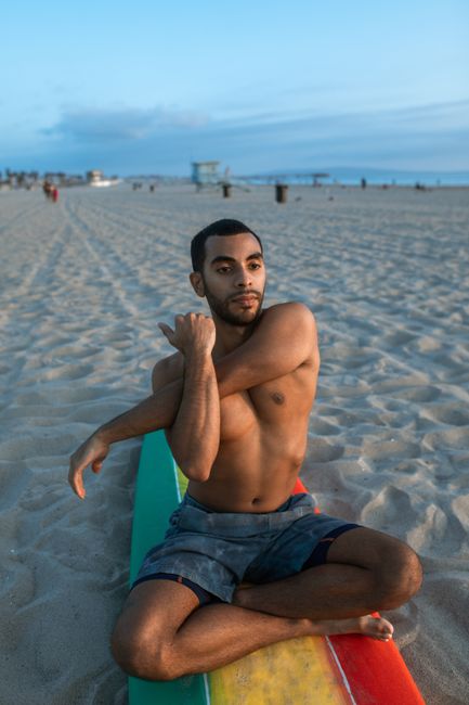 Shirtless man stretches on a colorful surfboard on a sandy beach at sunset.
