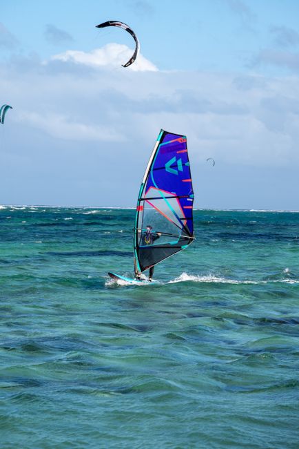 A thrilling windsurfing experience on clear blue ocean waters with kite surfers in the background.