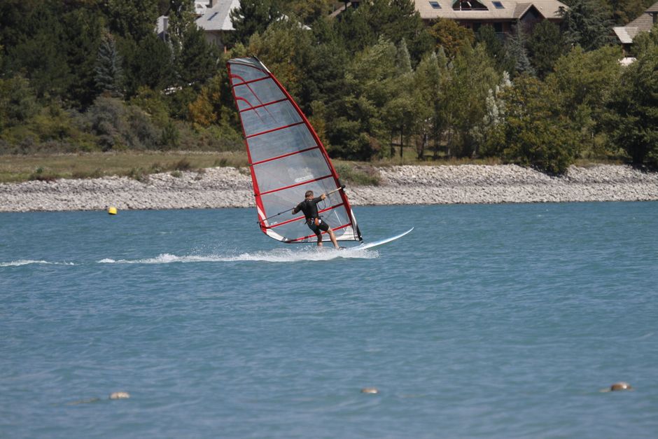 A person windsurfing on a clear lake with lush greenery in the background.
