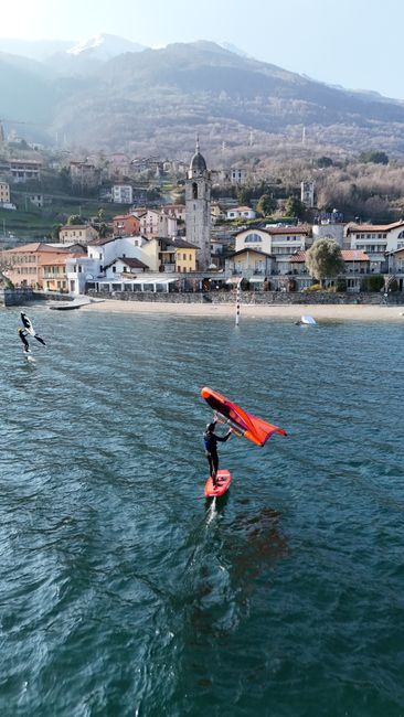 Wing surfer skims across Lake Como with Gravedona's scenic town and mountains in the background.