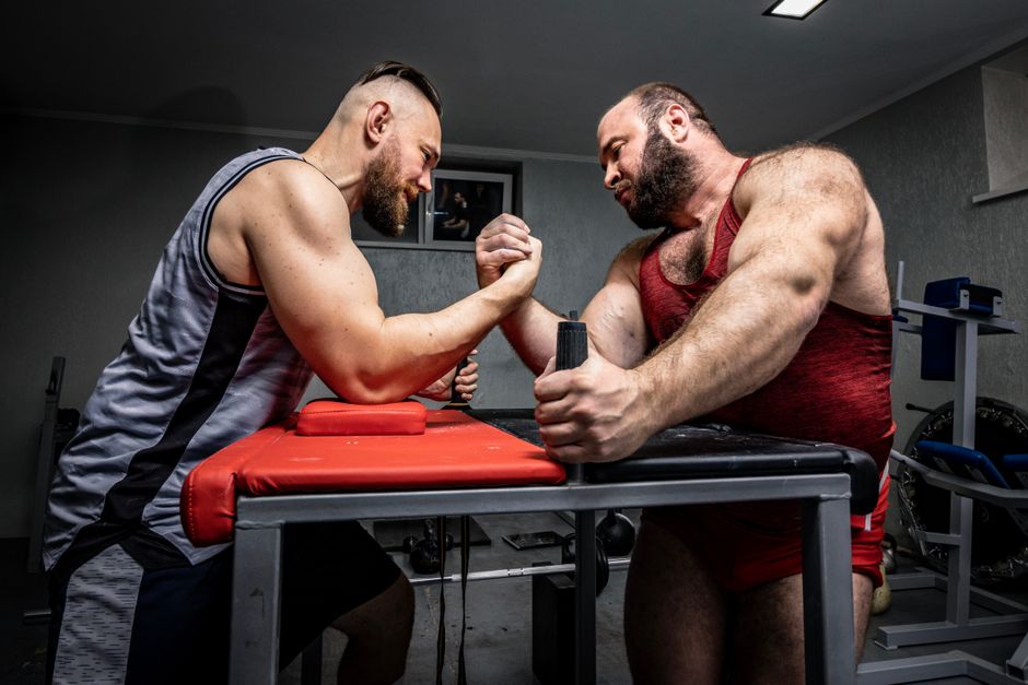 Two muscular men engaged in an intense arm wrestling match indoors.