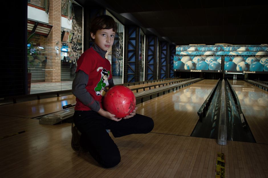 A young boy kneels holding a red bowling ball in a modern bowling alley scene.