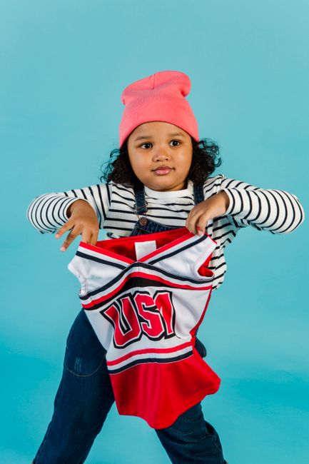 Cheerful African American girl wearing hat looking away while standing on blue background with cheerleader uniform in hands in studio