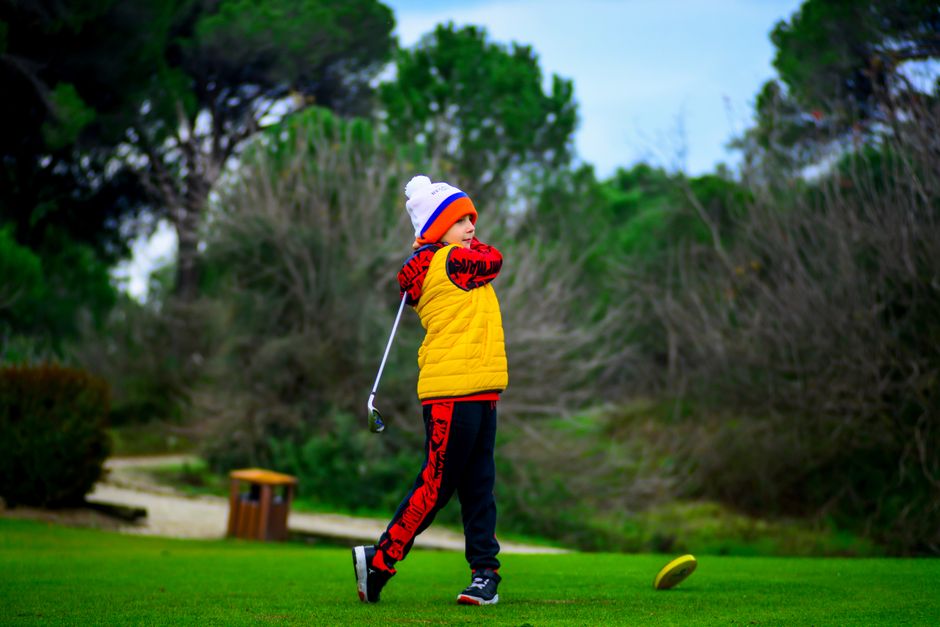 A child golfer in winter clothing swings a club on a lush green golf course.