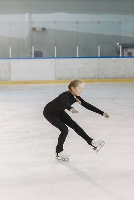 A young girl in black attire practicing figure skating in an indoor ice skating rink.