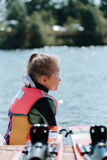 A young girl in a colorful life jacket sits by the water, ready for a wakeboarding adventure.